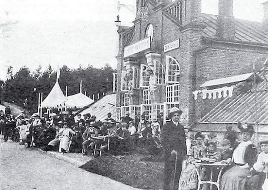 Crowds seated at outdoor tables beside a brick pavilion with awnings and a marquee tent, early 1900s