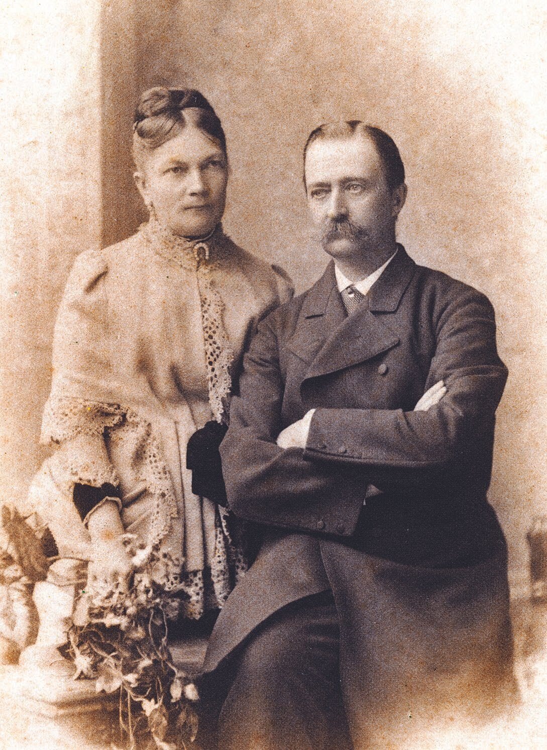 Sepia studio portrait of a seated moustached man with arms crossed beside a woman in lace dress