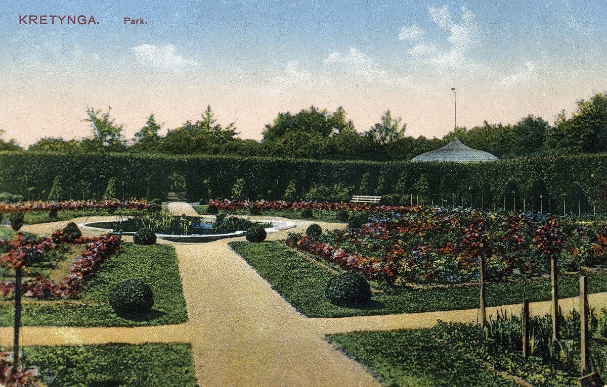 Hand-coloured postcard of a formal park with hedges, flower beds, clipped shrubs and a round pool