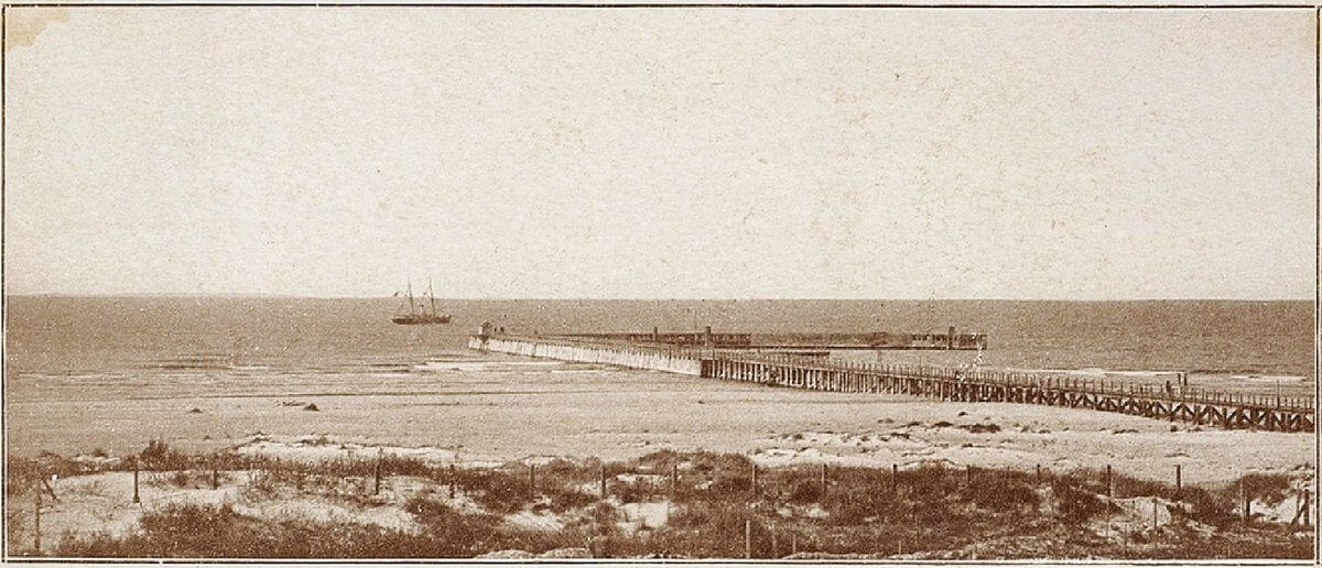 Sepia photograph of a long wooden pier stretching into the sea from a sandy dune shore, with a steamship offshore