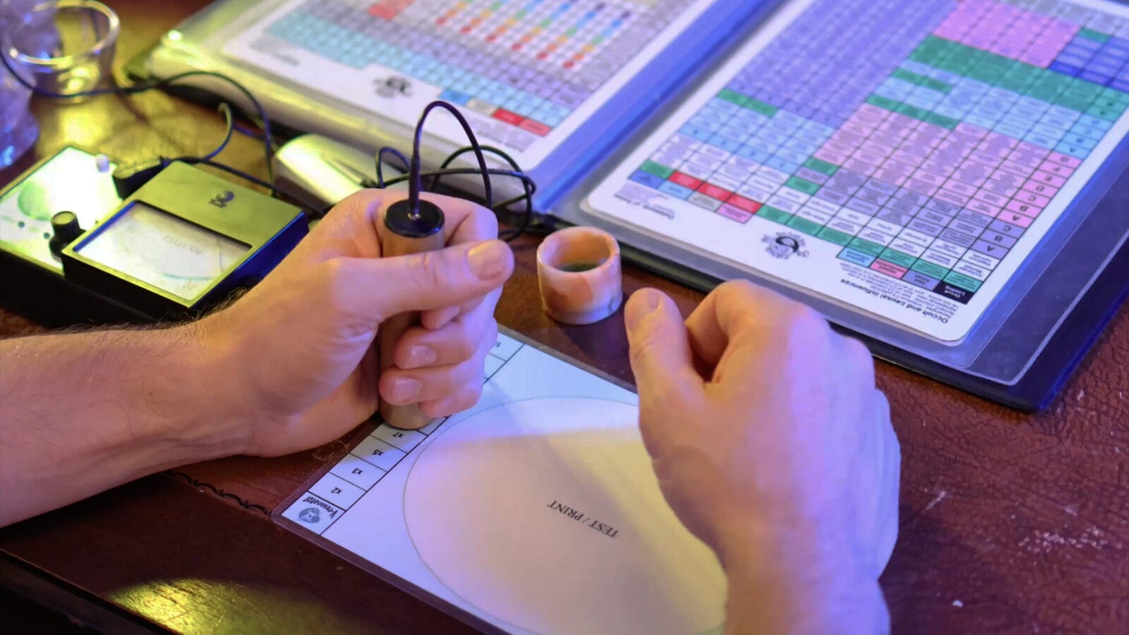 Radionics Practice With Rate Charts Hands holding a pendulum-like stick over a printed dial chart beside reference rate tables on a wooden desk