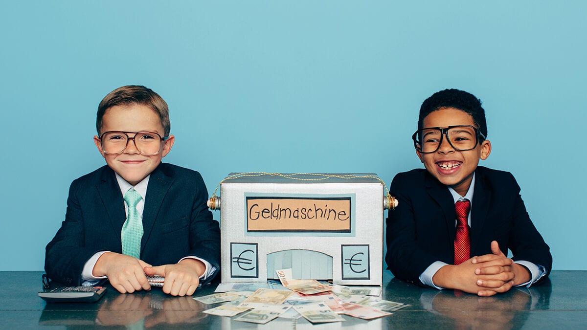 Two boys in suits and glasses smile beside a cardboard money machine spilling euro notes
